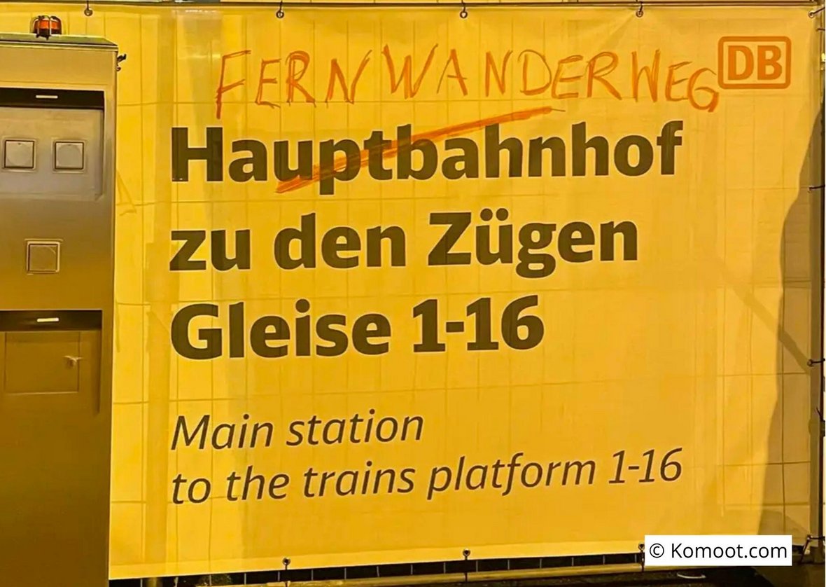 Fotografie eines Bauzaunschilds "Hauptbahnhof zu den Zügen Gleise 1-16 / Main station to the trains platform 1-16" auf dem das Wort "Hauptbahnhof" mit Grafitti durchgestrichen und durch "Fernwanderweg" ersetzt wurde